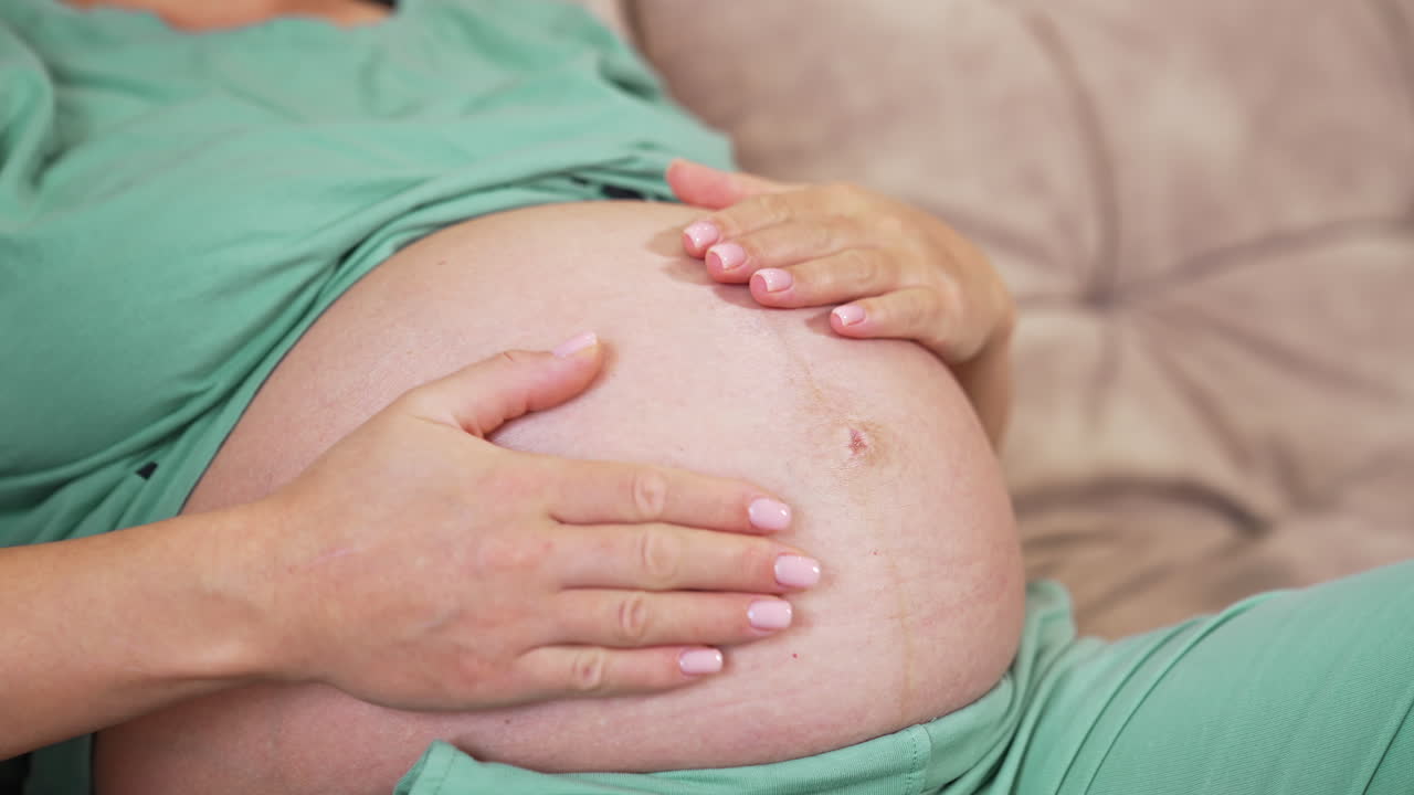 Bare stomach of a pregnant woman. Pregnant woman stroking her belly while laying on a sofa at home. Close-up. Pregnancy concept.