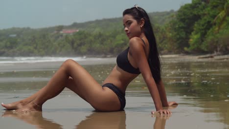 Amazing-young-woman-sits-in-the-sand-at-the-beach-in-a-black-bikini-with-hair-flowing-in-the-wind