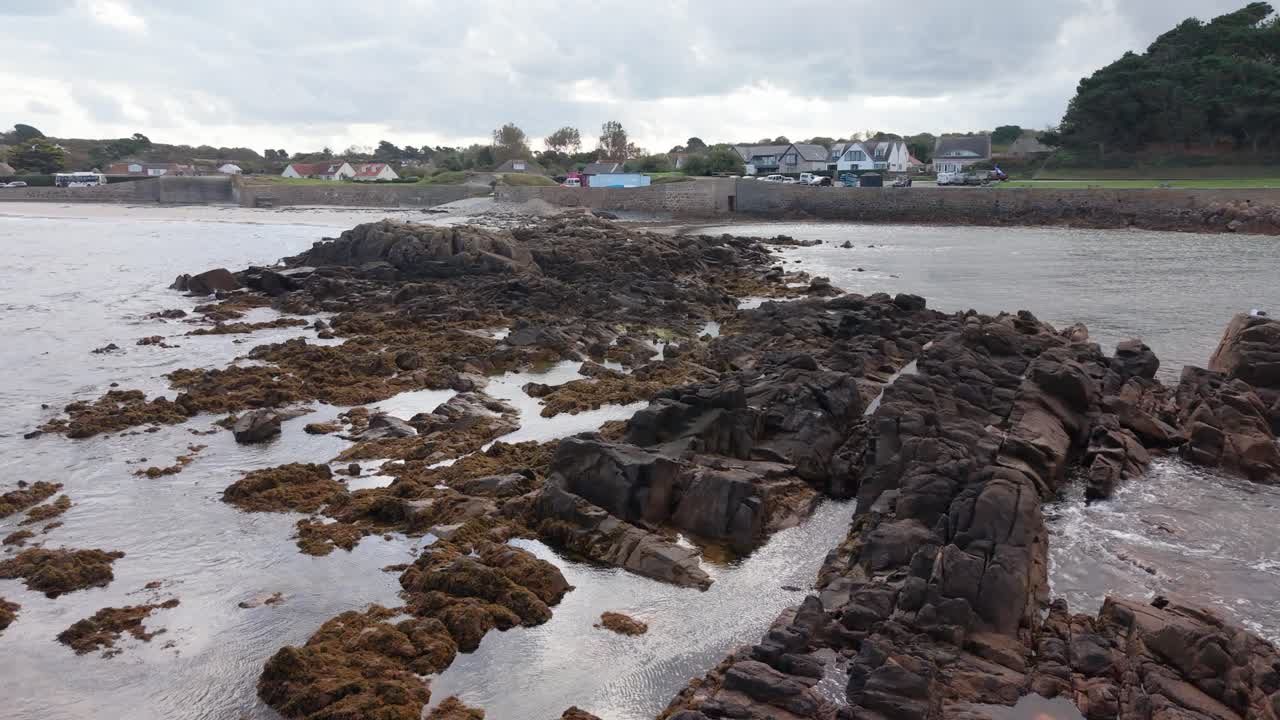 Slow Low forward flight over rocky outcrops from seaward heading towards the shoreline of sandy beach with seabirds taking flight on bright but cloudy day with interesting sky