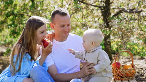 beautiful caucasian family sitting in the garden. mom shows the apple to her cute little baby.