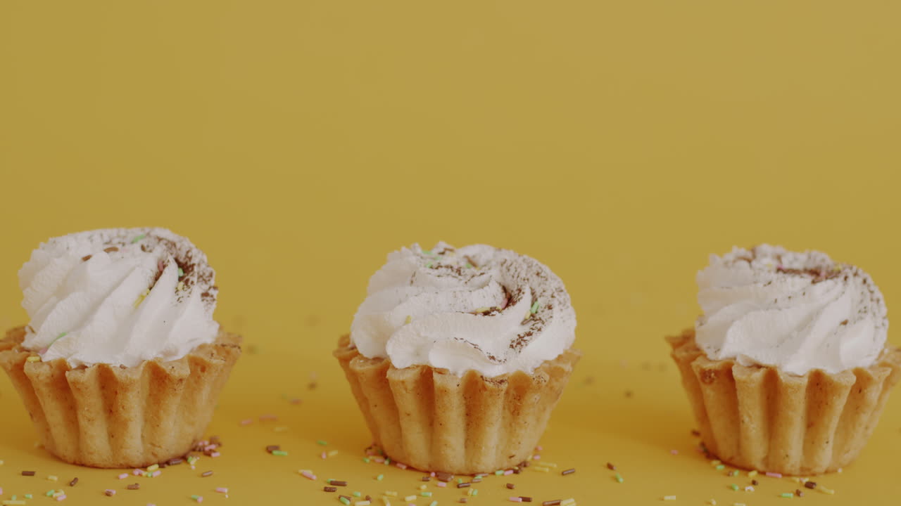 Three small cakes with whipped cream and sprinkles on a yellow background