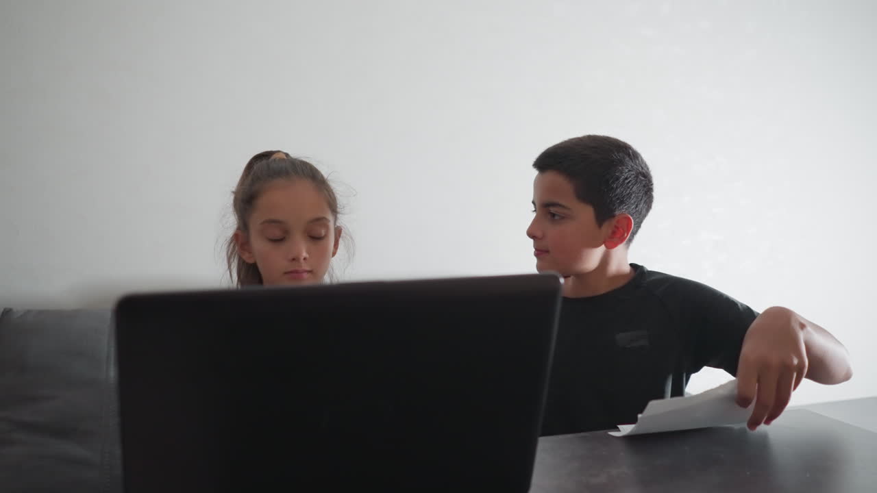 Una chica concentrada escribe en un portátil mientras su hermano, que está escribiendo, intenta llamar su atención mostrándole su trabajo. Ella mira brevemente su trabajo y le anima con una cálida sonrisa mientras él sigue escribiendo.