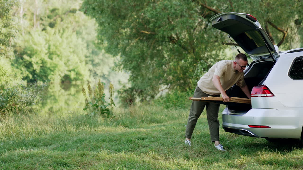 Man comes up to the car trunk. Man takes out the folding chair and puts it on the grass. Rest in nature.