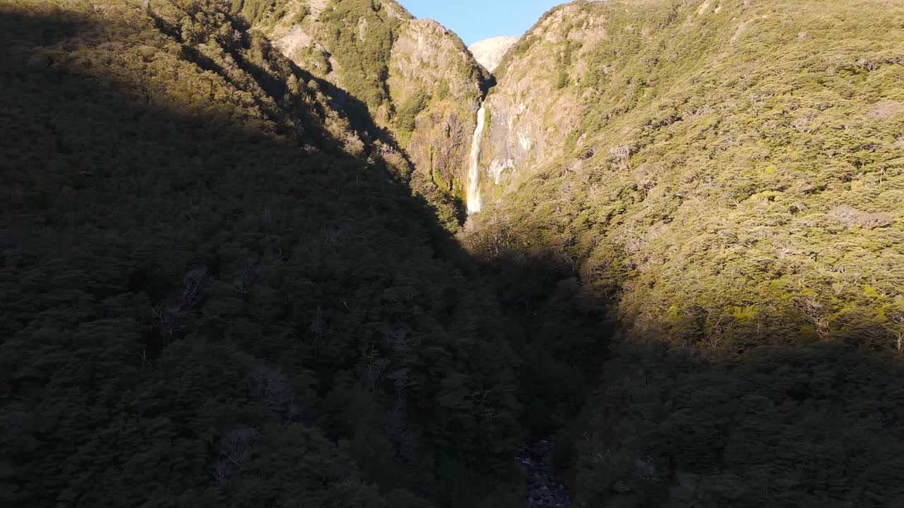 Drone rising over Devil’s Punchbowl falls in Arthur’s pass, New Zealand, sunset light on dense forest and cascading waterfall in alpine terrain.