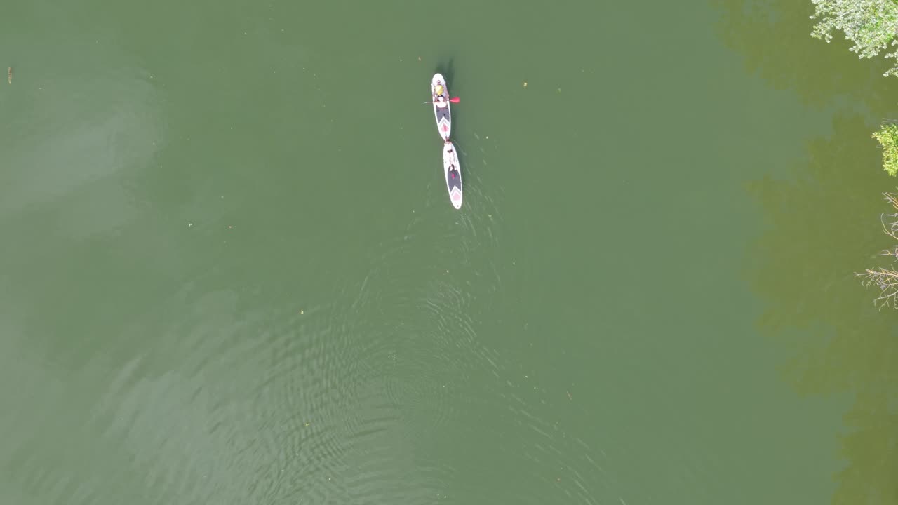 A top-down aerial view of two individuals paddleboarding side by side on tranquil green water. Their boards create gentle ripples as they move, surrounded by natural reflections and serene stillness