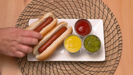 top down view of a hand placing hot dogs on a plate beside ketchup, mustard, and relish