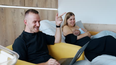 joyful caucasian couple with newborn. man works at laptop and his wife breastfeeding a baby.