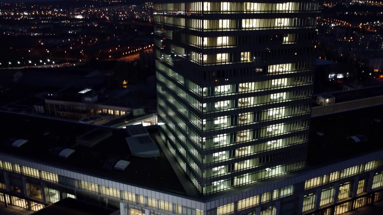 Torre de Escritórios Iluminada à Noite Exibindo Fachada de Vidro Iluminada e Janelas Brilhantes, Vista Aérea Sobre Praça Tranquila