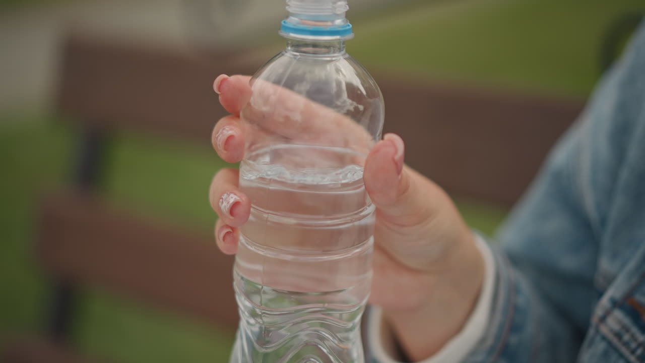 Nainen tarttuu vesipulloon puistonpenkillä. Lähikuva kädestä, joka pitää vesipulloa auringonvalossa. Yksityiskohtainen lähikuva naisen kädestä, joka pitää vesipulloa penkillä kukkakuvioisilla kynsillä.