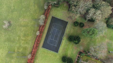 Vista-De-Drones-De-Arriba-Hacia-Abajo-De-La-Cancha-De-Tenis-De-Asfalto-Privada-En-Un-Jardín-Rodeado-De-árboles,-Césped-Y-Campos