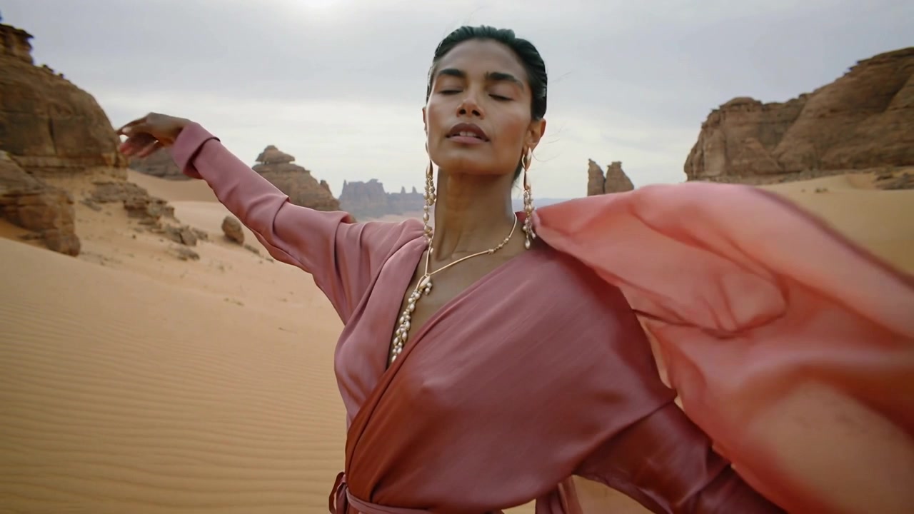 Turning wind-pushed female model unwrapping drape on sand dunes, showing pink dress and gold jewels