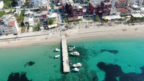 Drone-view-in-Albania-flying-over-a-beach-with-crystal-clear-blue-water-ocean,-buildings-on-the-harbor-and-a-wooden-walkway-on-a-sunny-day