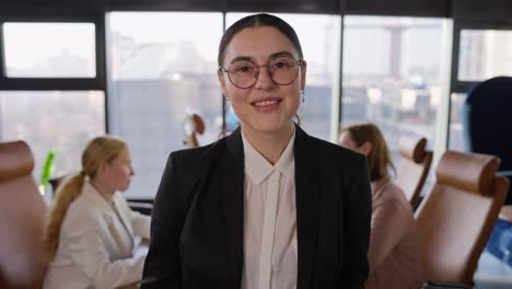 Portrait-of-a-confident-brunette-girl-in-a-business-suit-and-round-glasses-posing-near-a-wooden-table-and-her-colleagues-in-an-office-with-large-windows