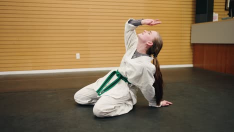 a young girl wearing a gi dobok sits on black rubber martial arts gym floor tired and exhausted from full day karate kung fu taekwondo training fake acts over exerted funny comical sports blooper