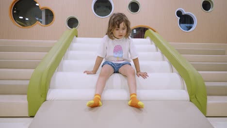 toddler sliding down indoor bumpy playground slide with excitement and fun in colorful play area, jumping up quickly with smile, low angle medium static, slow motion
