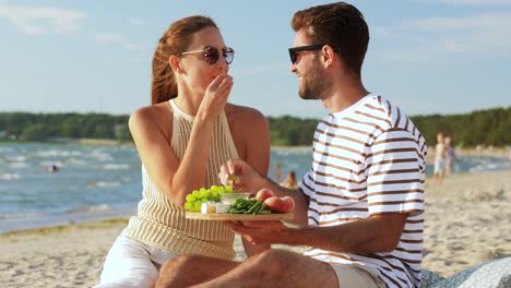 pareja feliz con la comida haciendo picnic en la playa. ocio, relaciones y personas concepto pareja feliz con los alimentos comiendo uvas y haciendo picnicen la playa