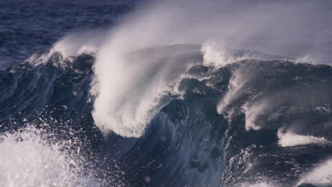 strong offshore winds blow spray from the top of a heavy barrelling wave as bursts of rainbow colour glimmer in the sunshine