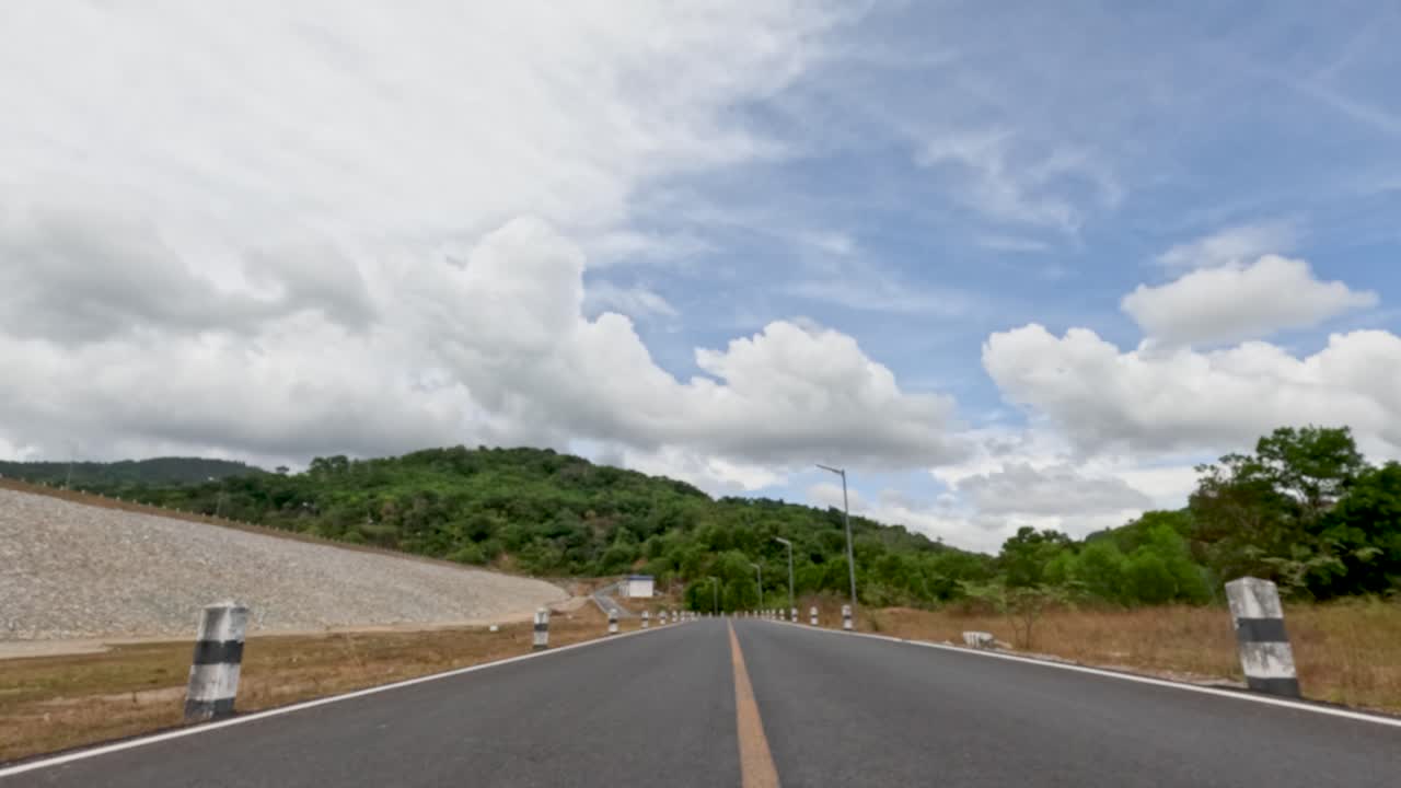 Car travels straight on empty rural road, daylight, wide angle, scenic hills, partly cloudy sky