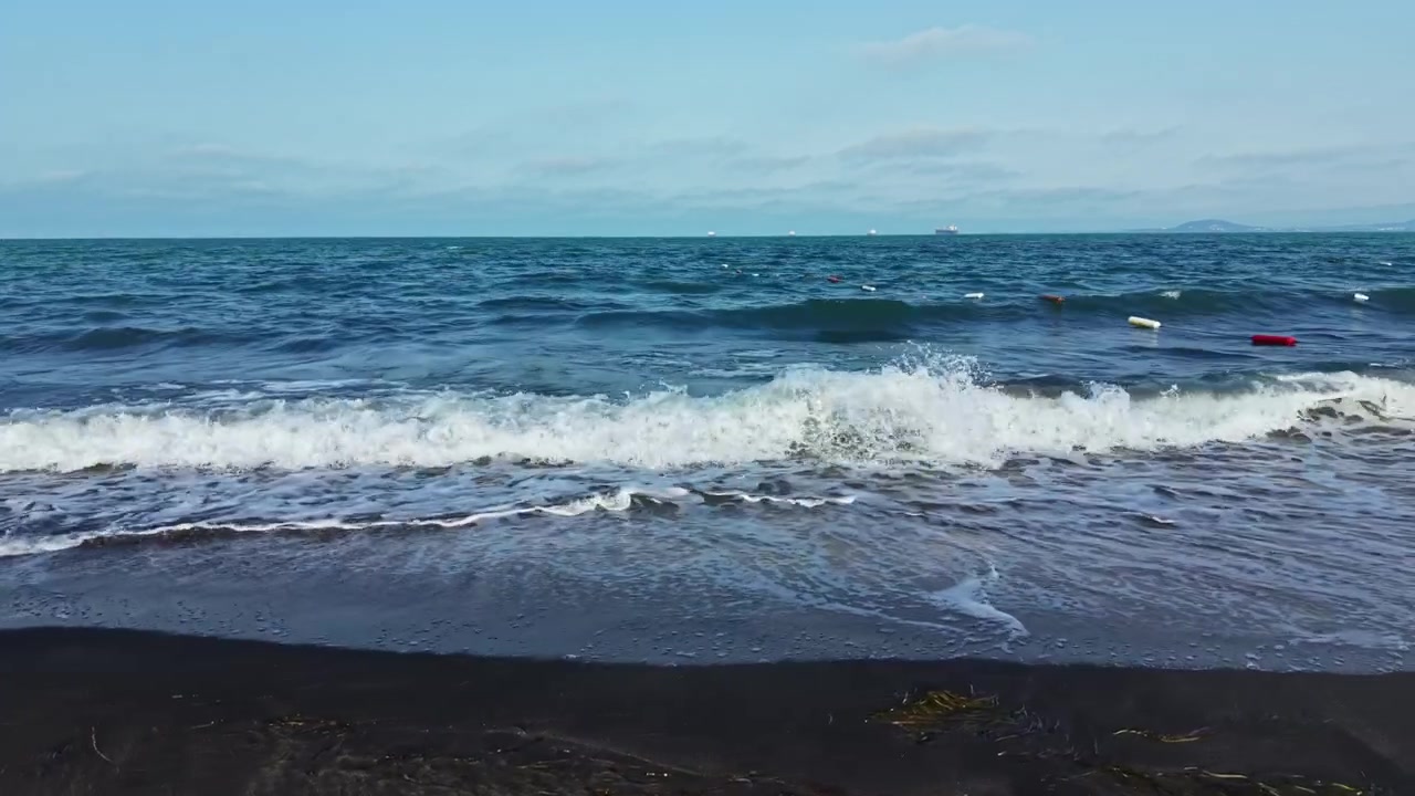Onde si infrangono su una spiaggia di sabbia scura con navi lontane e un cielo azzurro limpido sullo sfondo durante le ore diurne.