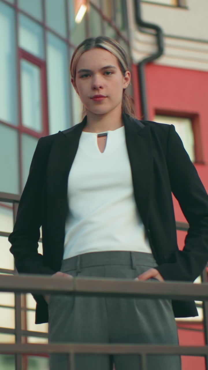 Elegant woman in black blazer and white blouse stands confidently with hands in pockets in front of residential building, iron railing and street light visible, tree in background