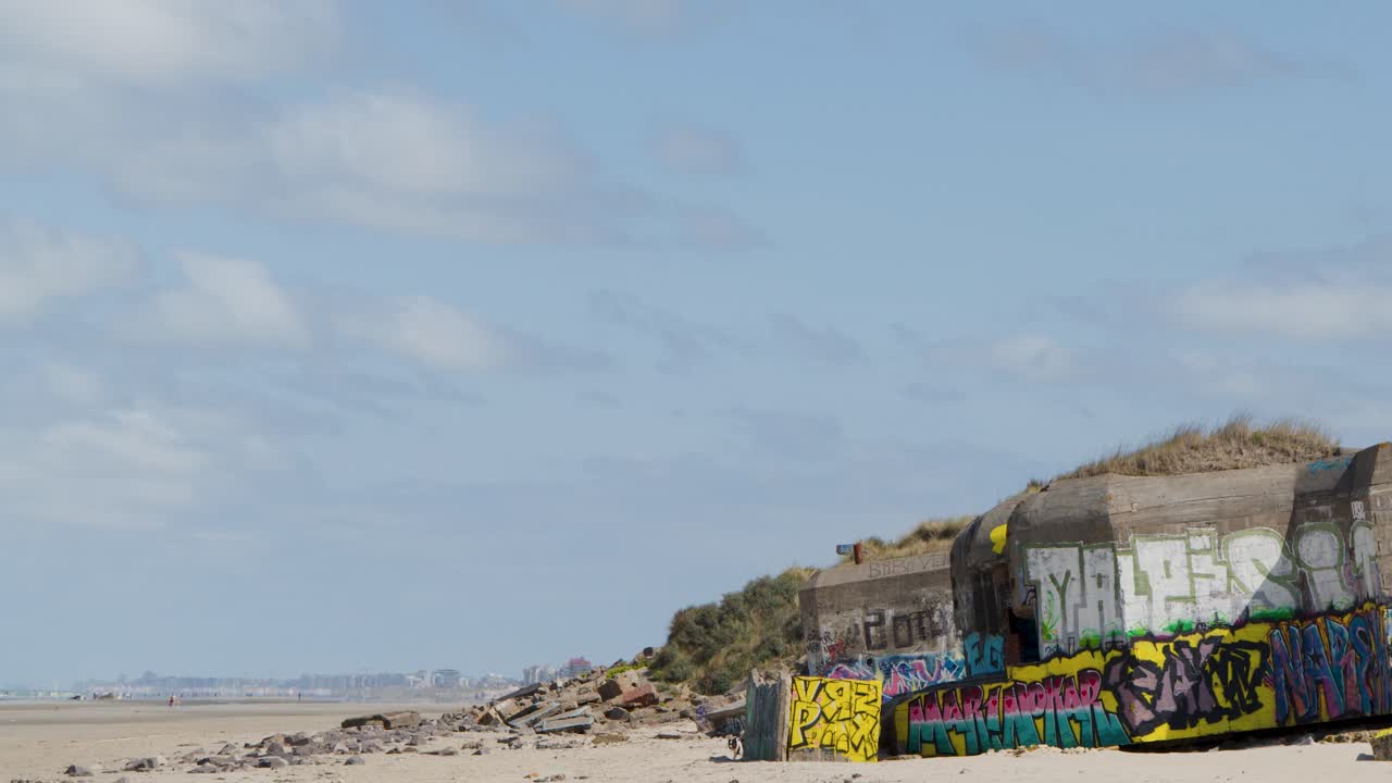 Wide shot of colorful graffiti-covered bunker ruins on sandy beach under bright daylight, clear sky
