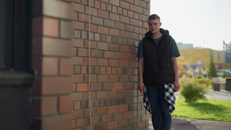 man walks slowly toward brick building wall and prepares to light cigarette using lighter while casually dressed in jeans and vest, with urban architecture and soft greenery blurred in background