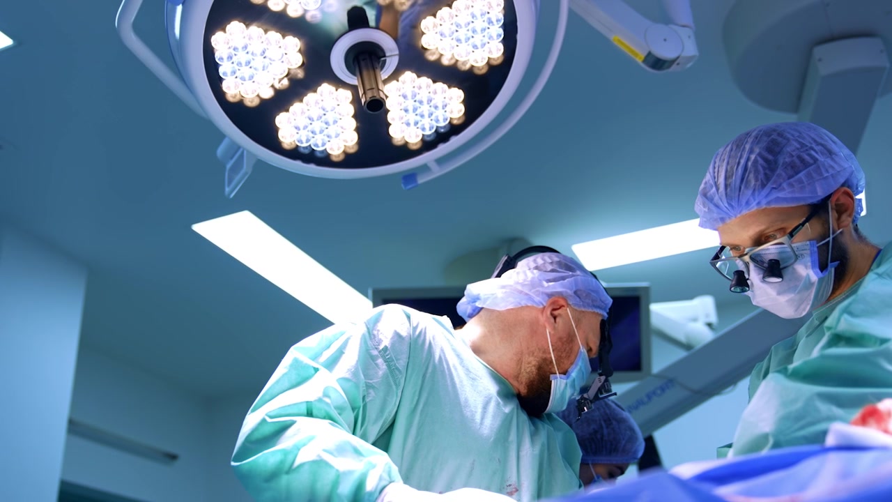 Doctores barbudos con lupas quirúrgicas cooperan en la cirugía en un hospital moderno. Equipo médico trabajando bajo una lámpara redonda. Vista en ángulo bajo.