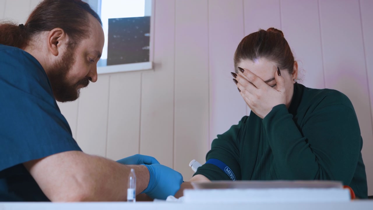 Paramedico che consola una donna in difficoltà durante una visita domiciliare, medico in divisa blu scuro con guanti offre una benda mentre è seduto al tavolo della cucina, luce diurna soffusa attraverso la finestra, supporto emotivo e valutazione.
