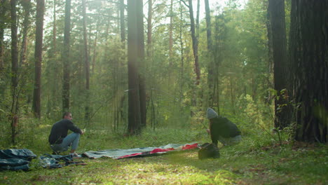 young man squats holding flexible tent pole while woman balances other end and lifts tent fabric during setup in lush forest, sunlight glows gently through trees, illuminating their teamwork