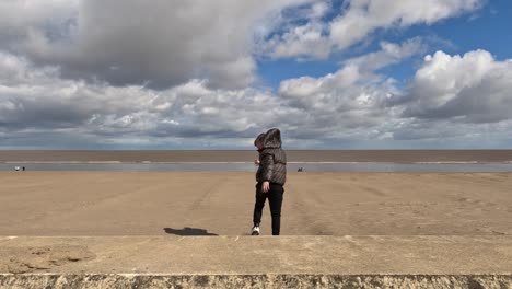 Young-boy-walking-along-a-wall-on-the-seafront-by-the-beach-on-a-stormy-sunny-day
