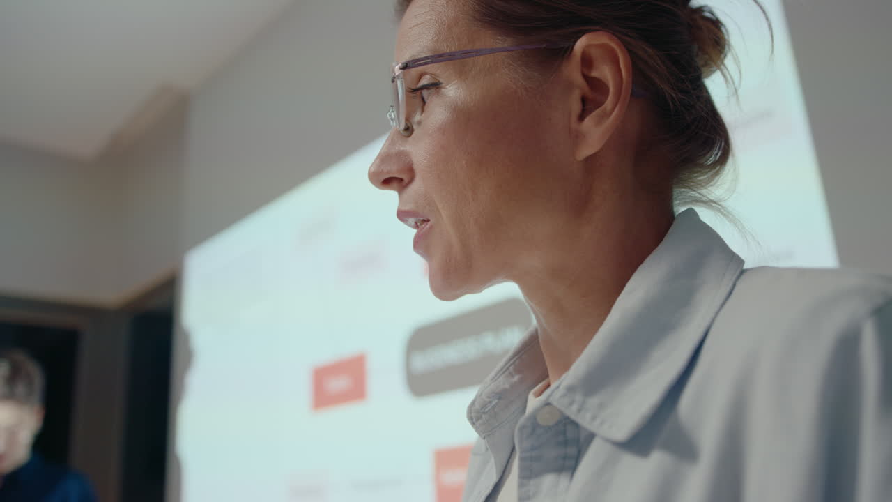 Businesswoman reading notes on paper cards and giving presentation during corporate meeting, projection screen and female colleague in the background. Close-up shot