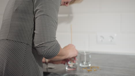 woman in plaid gown brings glass cup forward placing something inside with wall tiles and electric outlet visible in background, artistic project in progress, creative activity on kitchen countertop
