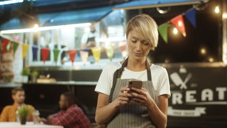 Young-Blonde-Woman-With-Short-Hair-In-Hair-Taping-And-Texting-Message-On-Smartphone-At-Festive-Food-Track-At-Night