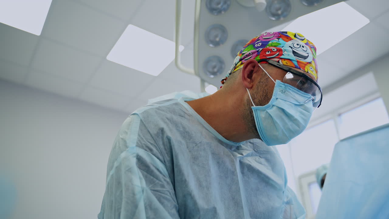 Adult male bearded surgeon in mask, glasses and bright cap working in surgery room. Low angle view from patient's point.