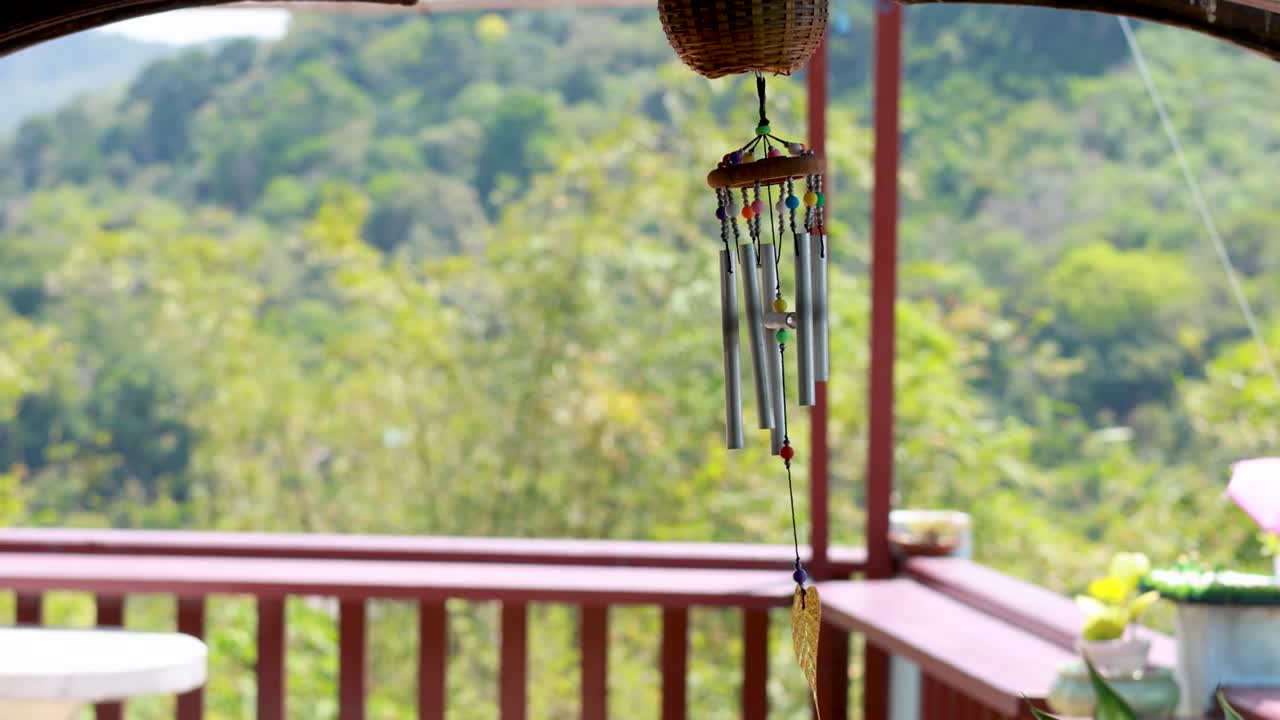 A wind chime gently sways on a porch overlooking lush greenery in Phuket, Thailand, under bright daylight