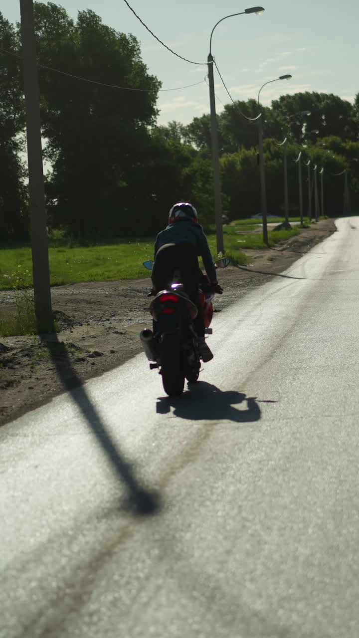vista trasera de una motociclista femenina de pie mientras monta una bicicleta eléctrica cerca de un camino fangoso, carretera vacía, el coche se acerca en la dirección opuesta, con árboles y edificios en el fondo