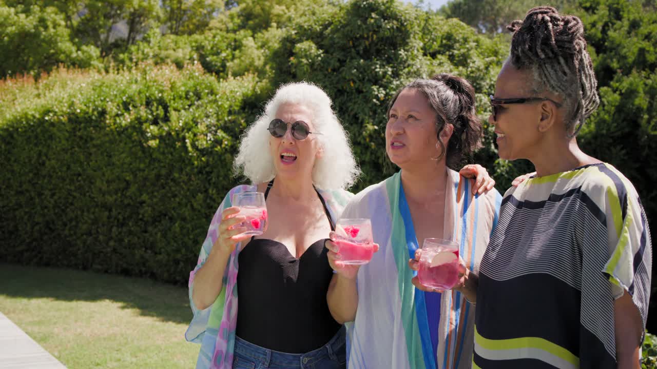 Un gruppo di donne mature con occhiali da sole si raduna a bordo piscina sotto il sole, chiacchierando e brindando con cocktail rosa.