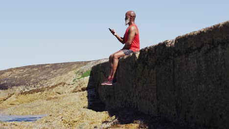 Älterer-Afroamerikanischer-Mann,-Der-Mit-Dem-Smartphone-Auf-Felsen-Am-Meer-Eine-Pause-Macht