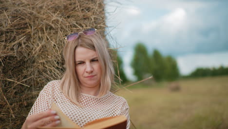 mujer con gafas de sol en la cabeza y uñas pintadas abre cuidadosamente el libro mientras se apoya en la bala de heno, un entorno al aire libre tranquilo con árboles borrosos en el fondo