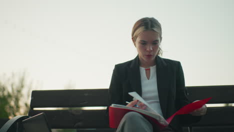 thoughtful ceo in formal wear seated alone on wooden bench outdoors reviewing business file with serious focus holding pen with laptop beside her in peaceful urban setting under bright sky