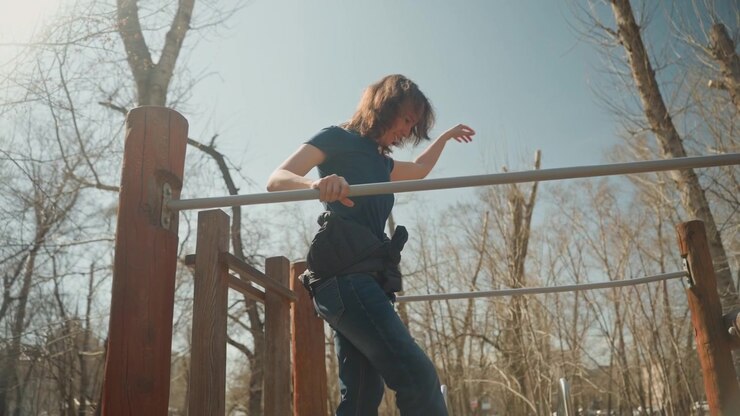 Close up of woman performing pull ups on outdoor exercise bar in daylight wearing casual clothing with sunlight illuminating scene and bare trees visible in background