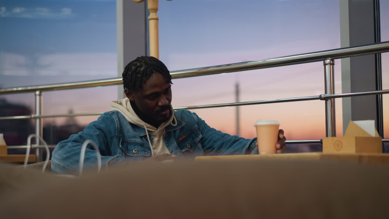 Young man in denim jacket and beige hoodie sitting in cozy cafe sipping coffee while looking into distance, surrounded by soft lighting, warm ambiance, large window view, and tranquil sunset reflections