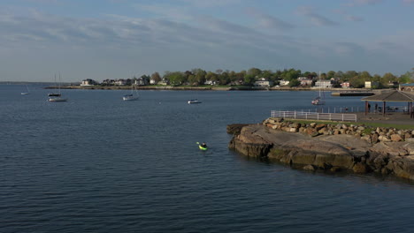 an aerial drone shot of a man paddling a kayak in calm waters by a park on a sunny day