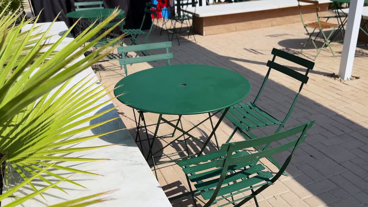 classic metal bistro table and chairs set outdoors on a sunny day.The scene captures strong shadows and a blurred palm frond foreground, emphasizing a timeless, European style cafe or garden aesthetic