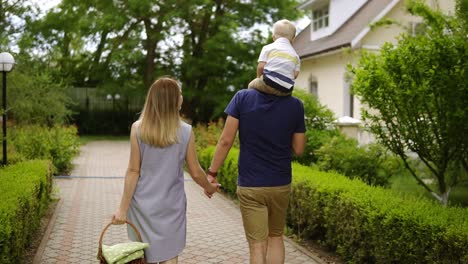 young family are walking otdoors at daytime. cottages territory. mother holding picnic basket on her hand. father carring a son on his shoulders. backside view