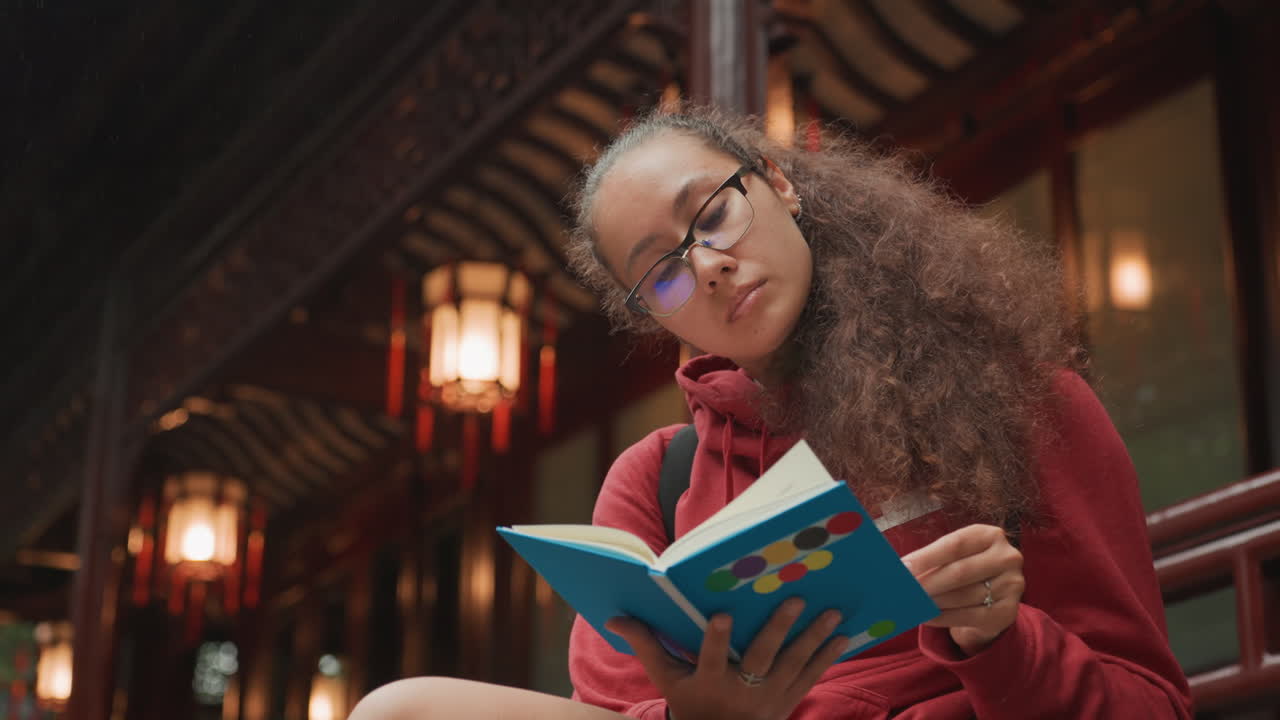 Jeune femme lisant sous des lanternes dans un pavillon couvert avec un éclairage chaleureux et un gros plan, esthétique d'étude nocturne avec des lunettes, cheveux bouclés, expression pensive et prise de notes créative