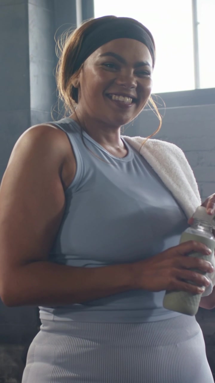 Video verticale: Donna di mezza età che beve, finendo l'allenamento in palestra, tenendo una bottiglia verde e un asciugamano.