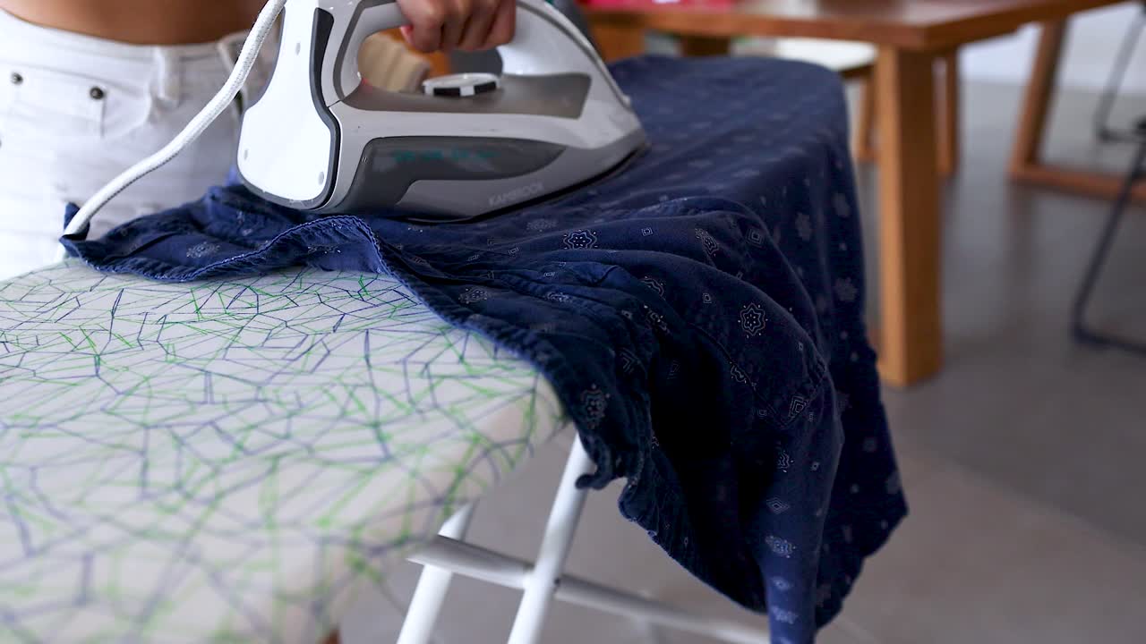 A person irons a blue garment on a patterned board in a bright, modern room