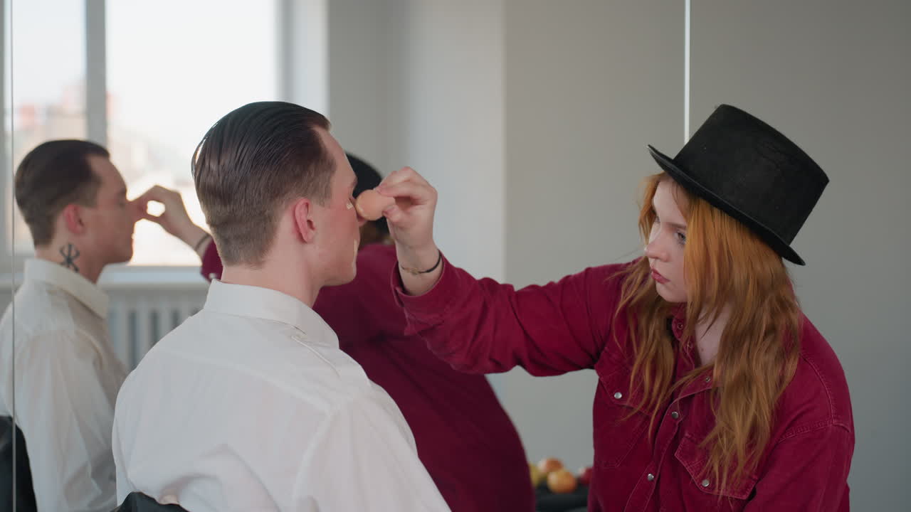 makeup artist applying foundation and concealer to male model face while mirror reflection reveals tribal neck tattoo in minimalist studio setting with natural light and intimate beauty session