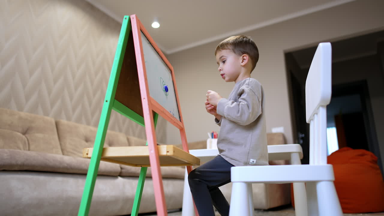 Kid drawing at home. A young boy is drawing with markers on a colorful easel in a cozy living room while sitting on a chair